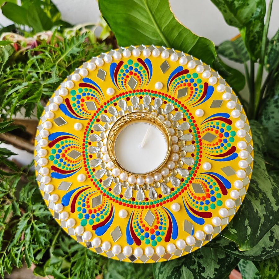 Round handmade candle plate painted in yellow with coloured dot art, pearls and mirror work, holding a lit tea light